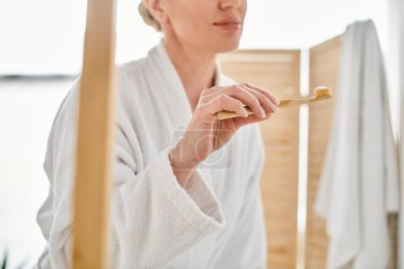 vue recadrée de la femme adulte en peignoir confortable mettre de la pâte dentaire sur sa brosse près du miroir