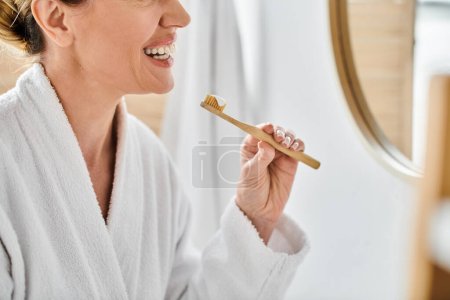 vista recortada de la alegre mujer adulta en albornoz poniendo pasta de dientes en su cepillo cerca del espejo
