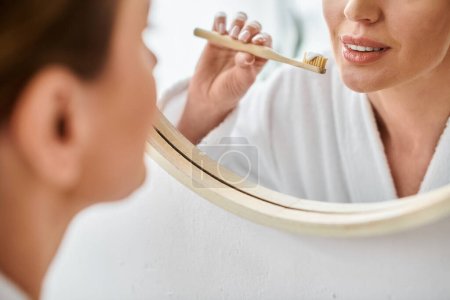 Ausgeschnittene Ansicht einer erwachsenen Frau im weißen Bademantel, die in der Nähe des Spiegels Zahnpasta auf ihre Bürste legt
