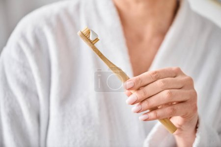 Ausgeschnittene Ansicht einer erwachsenen Frau im weißen Bademantel, die in der Nähe des Spiegels Zahnpasta auf ihre Bürste legt