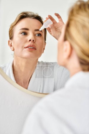 beautiful blonde woman with collected hair in bathrobe putting in eye drops in her bathroom