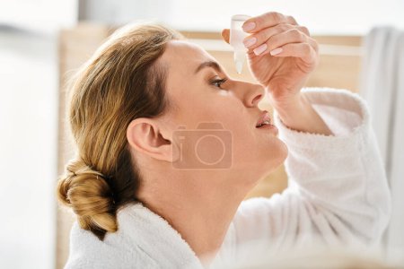 Photo for Attractive blonde woman with collected hair in bathrobe putting in eye drops in her bathroom - Royalty Free Image