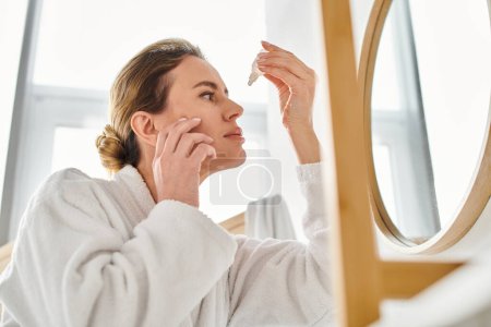 Photo for Appealing blonde woman with collected hair in bathrobe putting in eye drops in her bathroom - Royalty Free Image