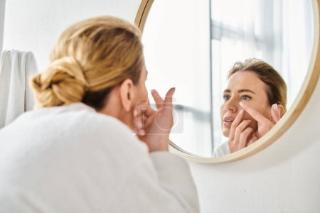 atractiva mujer en blanco cómodo albornoz usando sus lentes de contacto cerca del espejo en el baño