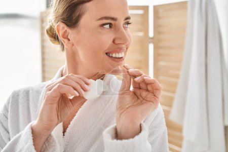 cheerful appealing woman in white bathrobe cleaning her teeth with dental floss in bathroom