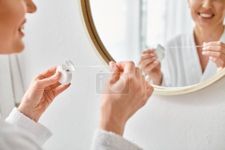 vista recortada de mujer adulta en acogedor albornoz limpiando sus dientes con hilo dental en el baño