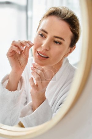 mujer atractiva alegre en albornoz blanco limpiando sus dientes con hilo dental en el baño