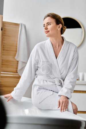 gut aussehende fröhliche Frau mit blonden Haaren im weißen kuscheligen Bademantel posiert neben ihrer Badewanne