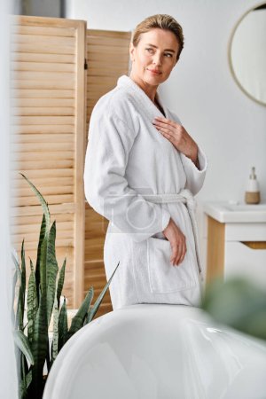 good looking happy woman with blonde hair in white cozy bathrobe posing next to her bathtub