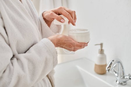 cropped view of adult woman in comfortable bathrobe holding face cream in her hands in bathroom