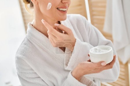 cropped view of cheerful woman in comfy bathrobe applying her face cream in bathroom at her home