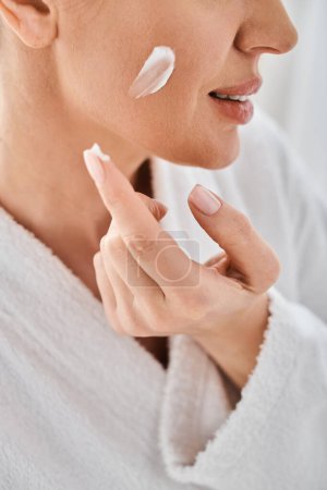 cropped view of cheerful woman in comfy bathrobe applying her face cream in bathroom at her home