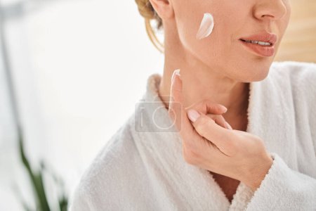 cropped view of adult woman in comfy bathrobe applying her face cream in bathroom at her home