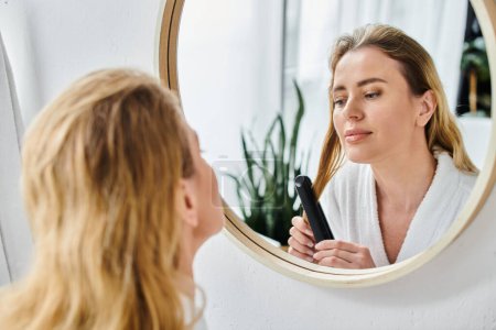 attractive blonde woman in cozy bathrobe using flat iron on her hair and looking at mirror