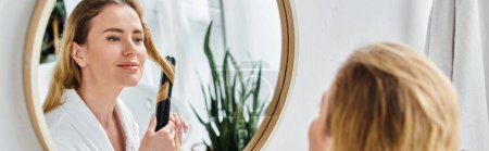 Téléchargez les photos : Joyeuse femme blonde attrayante en peignoir confortable en utilisant du fer plat sur ses cheveux pendant qu'elle est dans la salle de bain - en image libre de droit