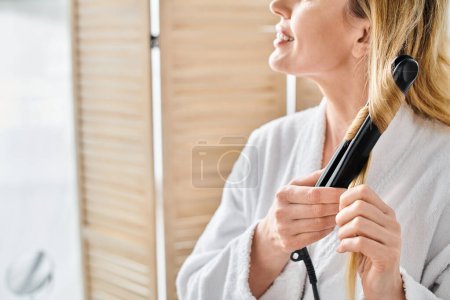 Téléchargez les photos : Vue recadrée de joyeuse femme blonde en peignoir confortable en utilisant du fer plat sur ses cheveux dans la salle de bain - en image libre de droit