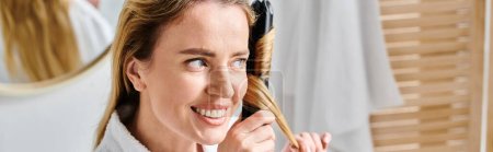 Téléchargez les photos : Joyeuse femme attrayante en peignoir confortable à l'aide de fer plat sur ses cheveux tandis que dans la salle de bain, bannière - en image libre de droit