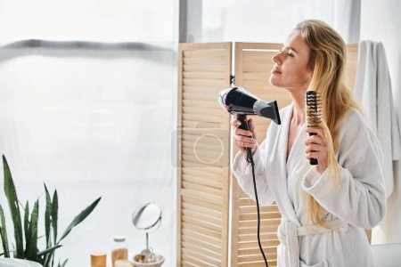 appealing blonde joyous woman in bathrobe using hair dryer and brush on her long hair at home
