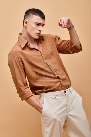 Retrato de moda de hombre guapo en camisa beige mirando hacia otro lado con la mano cerca de la cara sobre fondo beige
