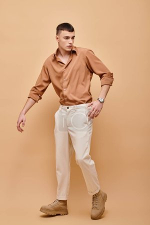 Full length image of young man in beige shirt, pants and boots posing on beige background