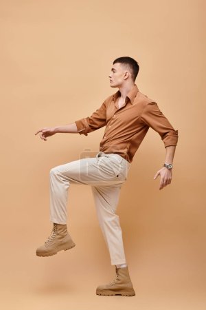 Téléchargez les photos : Photo latérale du jeune homme en chemise, pantalon et bottes beige posant sur fond beige - en image libre de droit