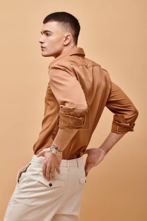 Back view portrait of stylish man in beige shirt with hand on pants on beige background