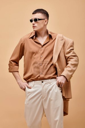 Eleganter Mann in beiger Jacke auf Schulter, Hemd, Hose und Sonnenbrille posiert auf beigem Hintergrund