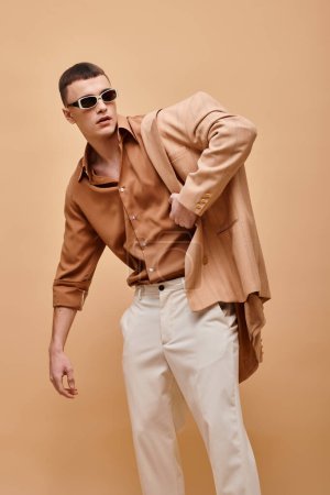 Confident man in beige jacket on shoulder, shirt, pants and sunglasses posing on beige background