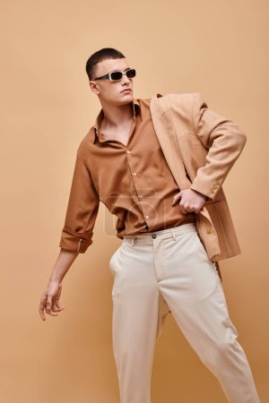 Hombre de moda en chaqueta beige en hombro, camisa, pantalones y gafas de sol sobre fondo beige, bandera