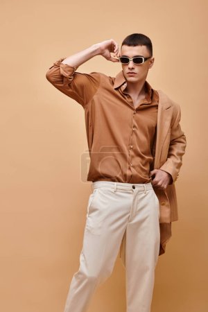 Classy man in beige jacket on shoulder, shirt, pants and sunglasses posing on beige background