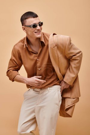 Smiling man in beige jacket on shoulder, shirt, pants and sunglasses on beige background