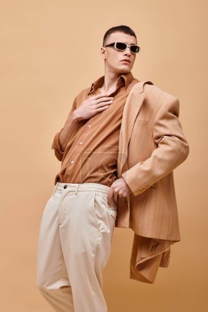 Fashion shot of man in beige jacket and shirt with sunglasses and hand near collar on beige backdrop