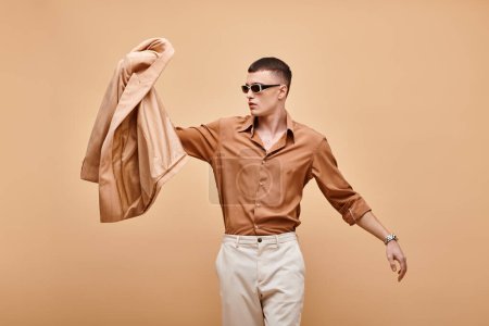 Fashion shot of man in beige shirt with sunglasses and holding jacket in hand on beige backdrop