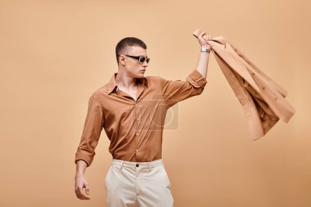 Fashion shot of man in beige shirt with sunglasses and throwing jacket away on beige backdrop
