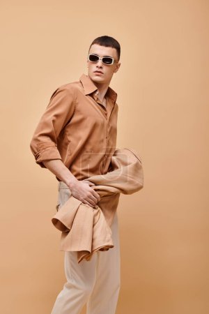 Fashion shot of man in beige shirt with sunglasses and holding jacket in hand on beige backdrop