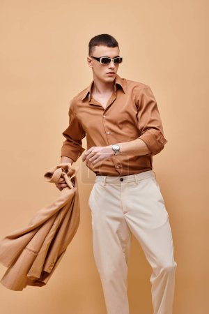 Portrait of man in beige jacket and shirt with sunglasses holding jacket in hand on beige backdrop