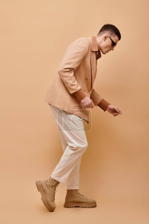 Téléchargez les photos : Image latérale de l'homme élégant en veste, chemise, pantalon et bottes beige posant sur fond beige - en image libre de droit