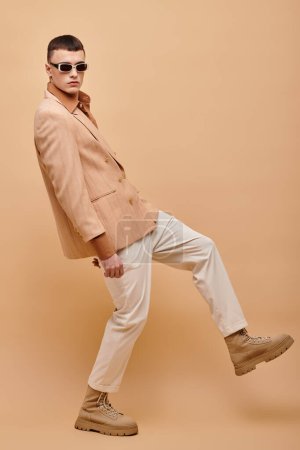 Retrato de hombre en chaqueta beige, camisa, pantalones y botas posando sobre fondo beige, estandarte