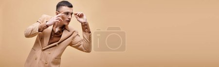 Handsome man in beige jacket touching glasses posing on peachy beige background, banner