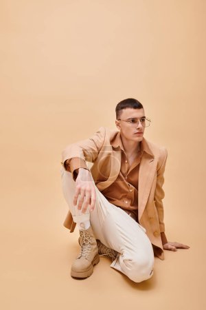 Foto de Hombre guapo en chaqueta beige y gafas sentadas sobre fondo beige melocotón - Imagen libre de derechos