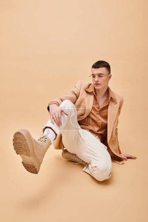 Foto de Hombre con estilo en chaqueta beige y gafas sentado y posando sobre fondo beige melocotón - Imagen libre de derechos