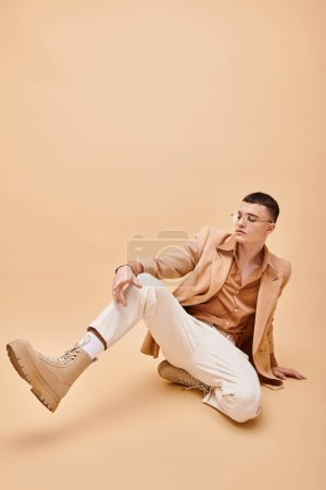 Hombre con estilo en los años 20 en chaqueta beige y gafas sentadas sobre fondo beige melocotón