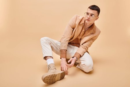 Téléchargez les photos : Young man in his 20s wearing beige jacket and glasses sitting on peachy beige background - en image libre de droit