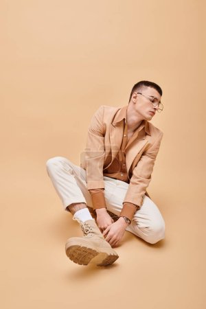 Handsome man in beige jacket and glasses sitting on peachy beige background
