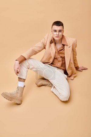 Stylish man in beige jacket and glasses sitting and posing on peachy beige background