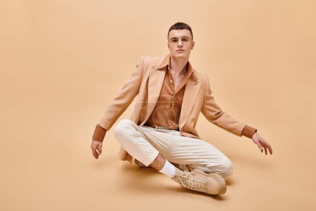 Foto de Hombre guapo en chaqueta beige y gafas sentadas en pose de loto sobre fondo beige melocotón - Imagen libre de derechos
