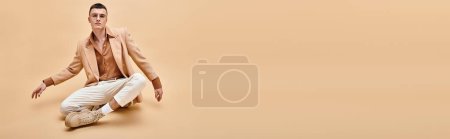 Handsome man in beige jacket and glasses sitting in lotus pose  on peachy beige background, banner