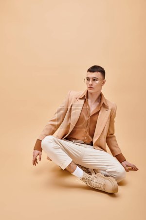 Fashion shot of man in beige jacket and glasses sitting in lotus pose  on peachy beige background
