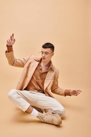 Photo for Handsome man in beige jacket and glasses sitting in lotus pose on peachy beige background - Royalty Free Image