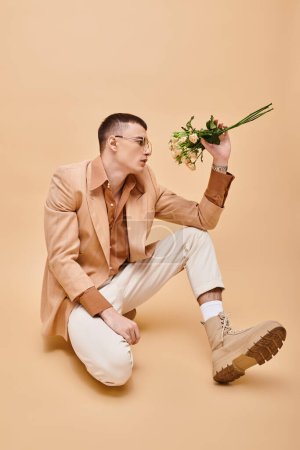 Téléchargez les photos : Portrait de mode d'homme en veste beige et lunettes assises avec des roses sur fond beige pêche - en image libre de droit